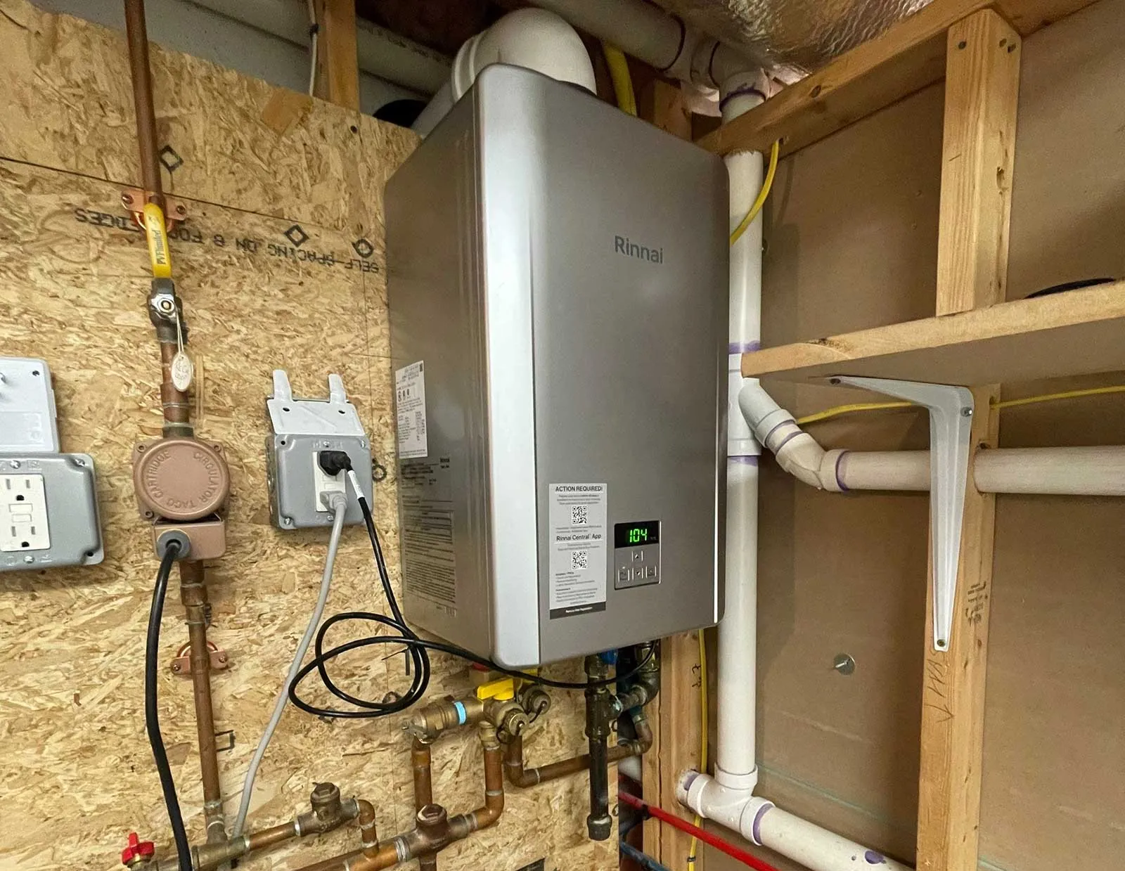 A gas tankless water heater installation on a plywood wall, with the copper gas line, white PVC vent pipes and a dedicated outlet